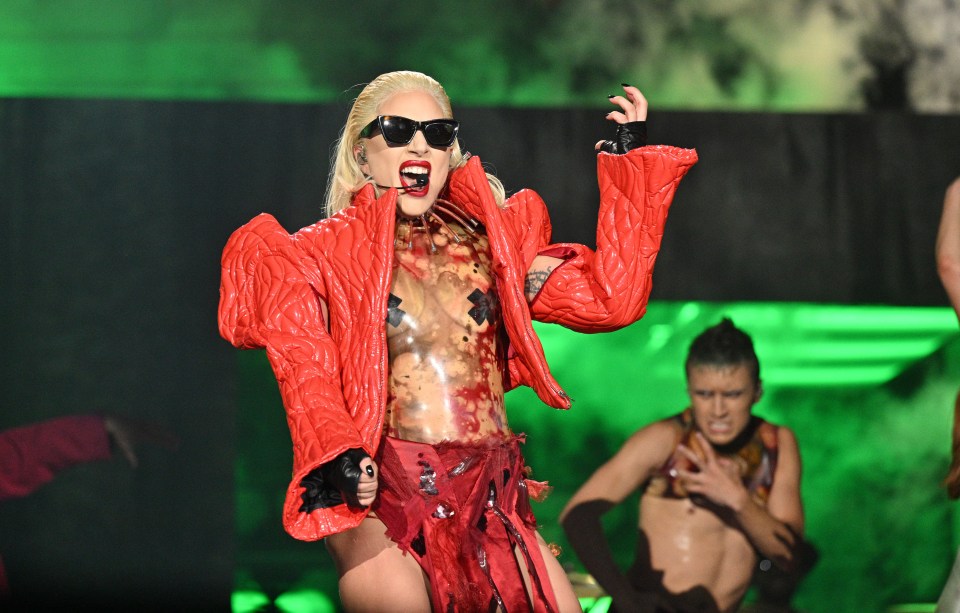 Lady Gaga performing on stage in a red jacket.