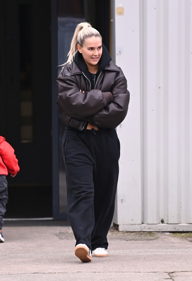 Molly Mae walking, smiling, wearing a brown leather jacket and black sweatpants.