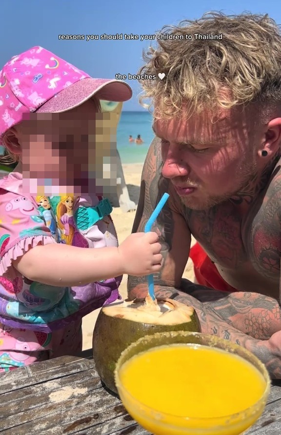 A toddler and a man on a beach drinking from a coconut.