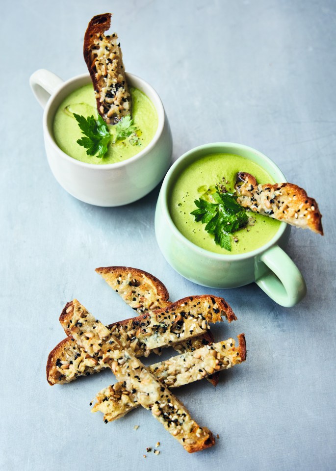 Two mugs of green soup topped with parsley and seed-topped breadsticks.