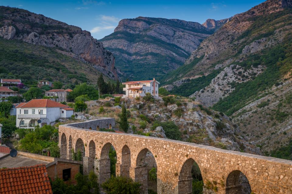 Ancient stone aqueduct in Bar, Montenegro, with houses and mountains in the background.