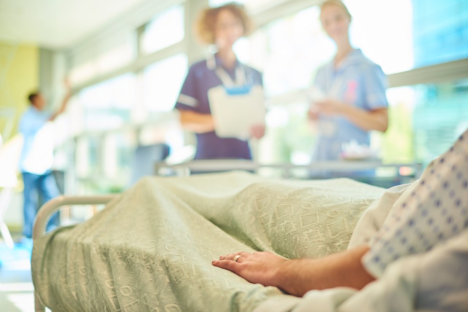 A patient's hand rests on a hospital bed, with blurred nurses in the background.