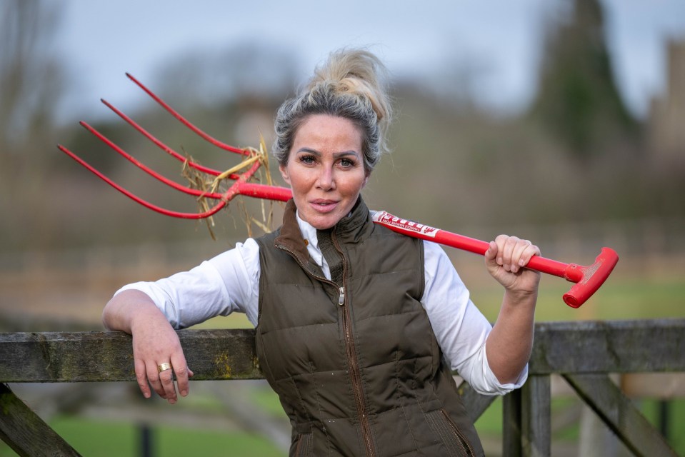 Woman with blonde hair, wearing a vest and white shirt, leans on a wooden fence with a pitchfork over her shoulder.