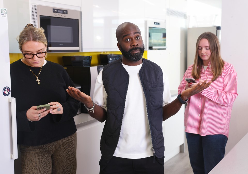 Three coworkers in an office kitchen, one looking perplexed as the others check their phones.