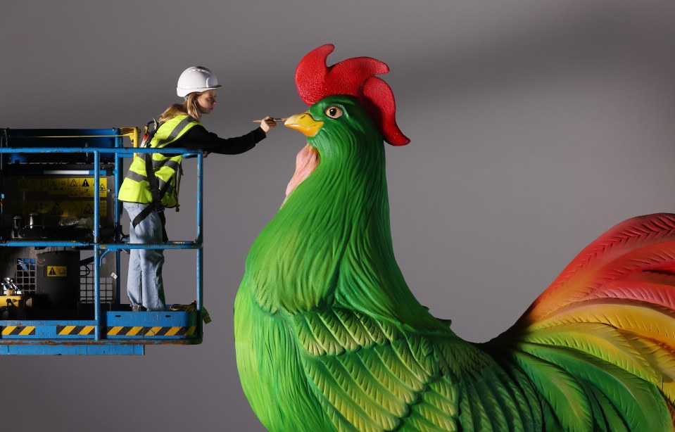 A worker in a lift painting a large rooster sculpture.