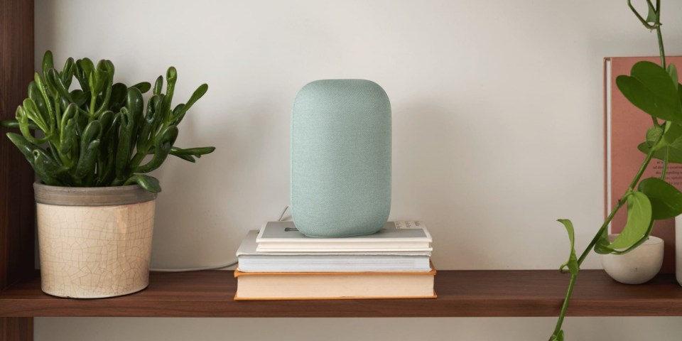 A light teal smart speaker sits on a stack of books on a shelf.