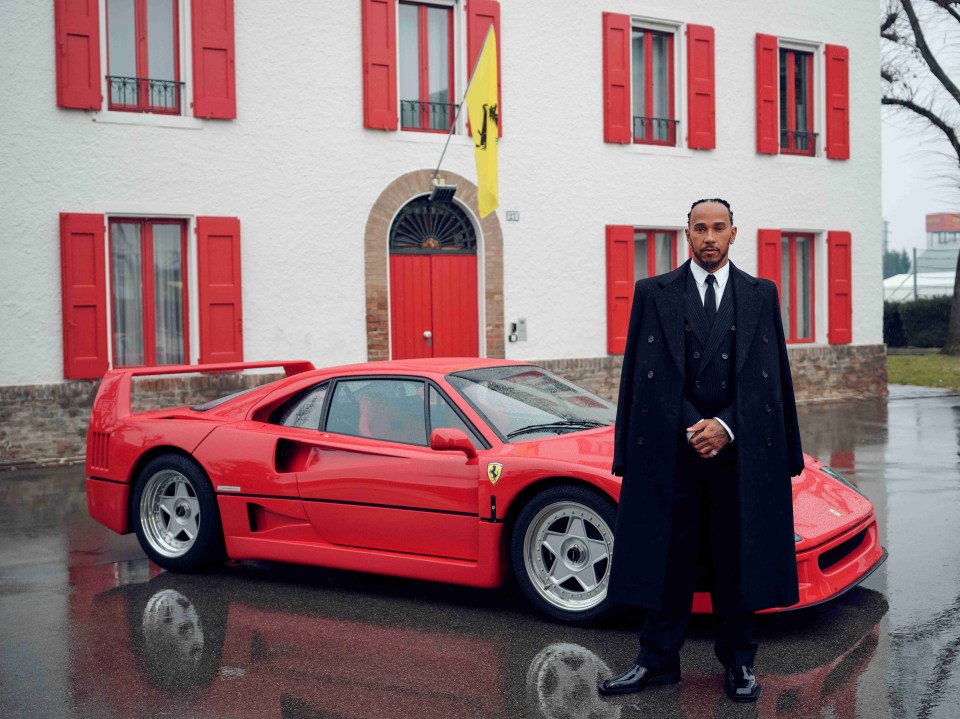 Lewis Hamilton in a black suit and overcoat, standing in front of a red Ferrari.