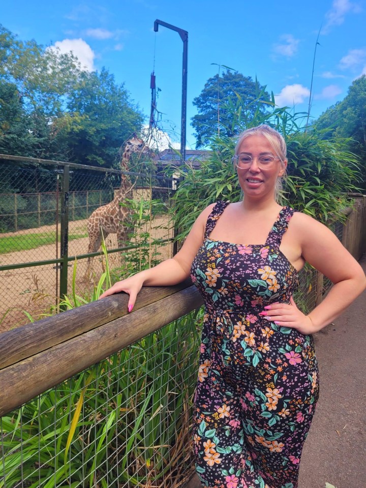 Woman in floral jumpsuit at a zoo, with a giraffe in the background.