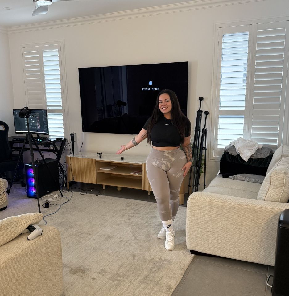 Woman standing in her living room, which includes a large TV, gaming setup, and beige couch.