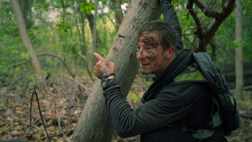 A mud-covered man points in a forest.