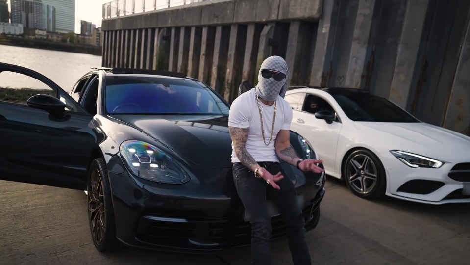 Man in a balaclava sitting on a black Porsche, with a white Mercedes-Benz behind him.