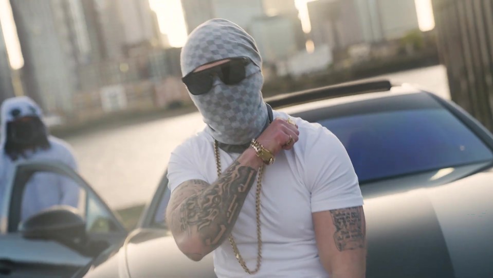 Screengrab of a man wearing a balaclava, sunglasses, and gold jewelry, standing by a car.