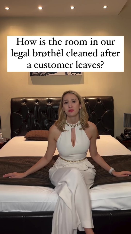 Woman in white jumpsuit sits on a bed in a legal brothel, answering a question about room cleaning.