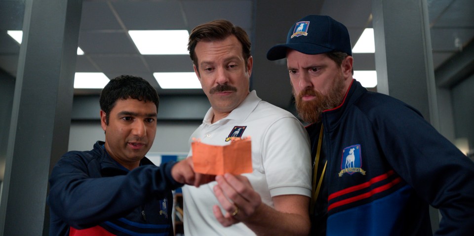 Still image from Ted Lasso showing Jason Sudeikis, Nick Mohammed, and Brendan Hunt looking at a piece of paper.