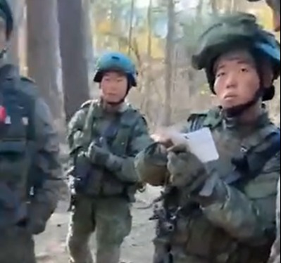 Three soldiers in camouflage uniforms in a wooded area; one soldier is holding a piece of paper.