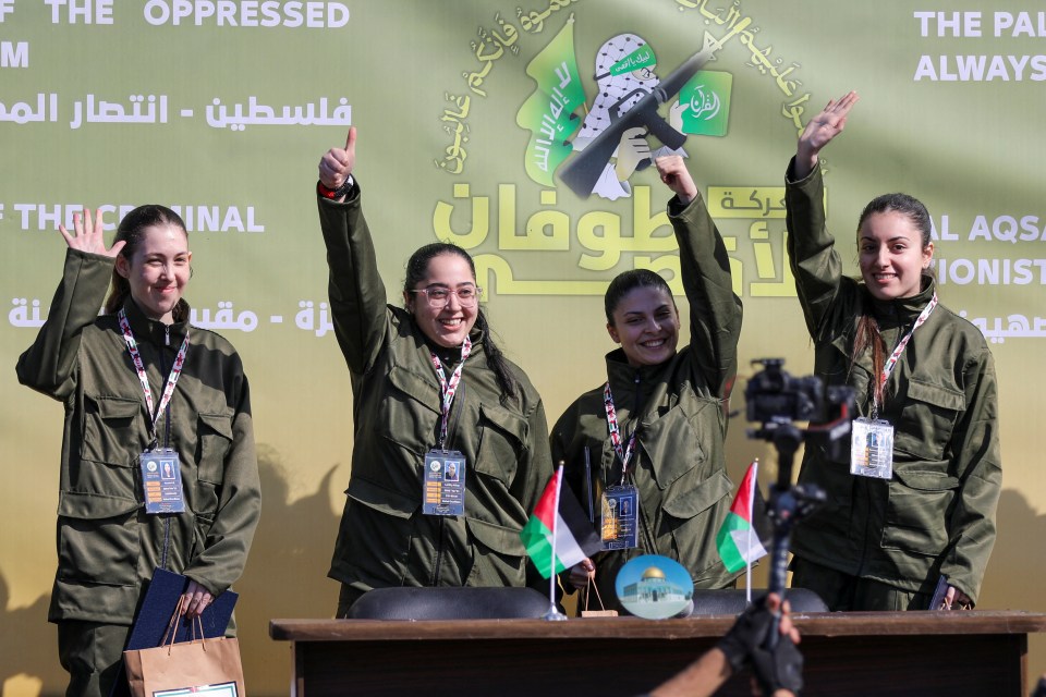 Four women in military-style uniforms stand behind a table, giving thumbs up.