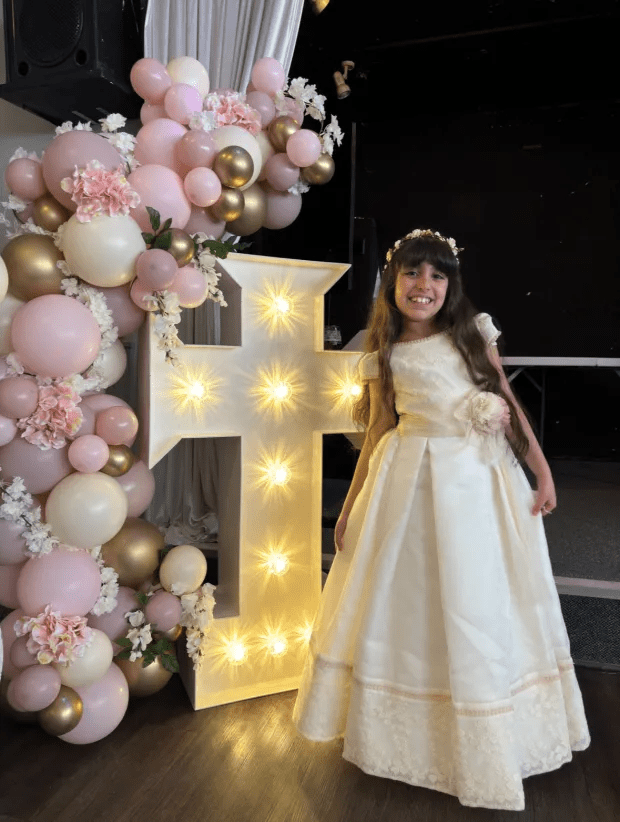 Girl in communion dress standing next to a lighted cross.