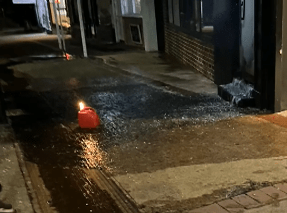 A burning fuel canister sits in a puddle of spilled fuel on a city street.