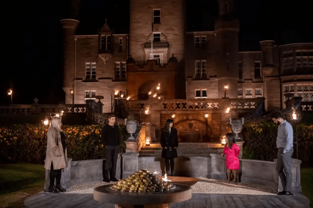 Five people stand outside a large castle at night, gathered around a fire.