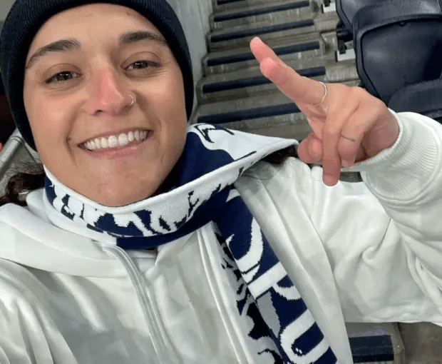 Smiling woman wearing a white hoodie and navy and white scarf, giving a peace sign.