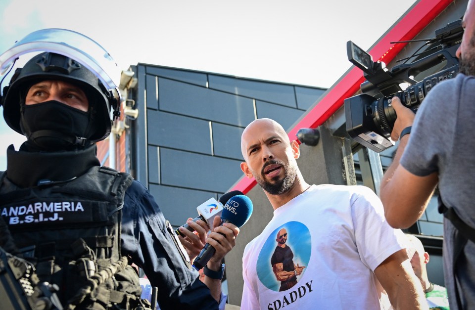 Man in white shirt with "SDaddy" and an illustration of a man printed on it speaks to reporters and police.