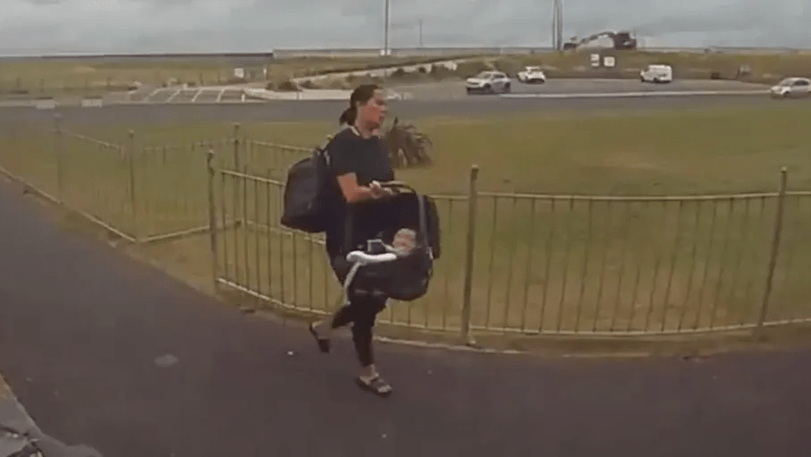 Woman carrying baby in car seat and backpack walks along path.