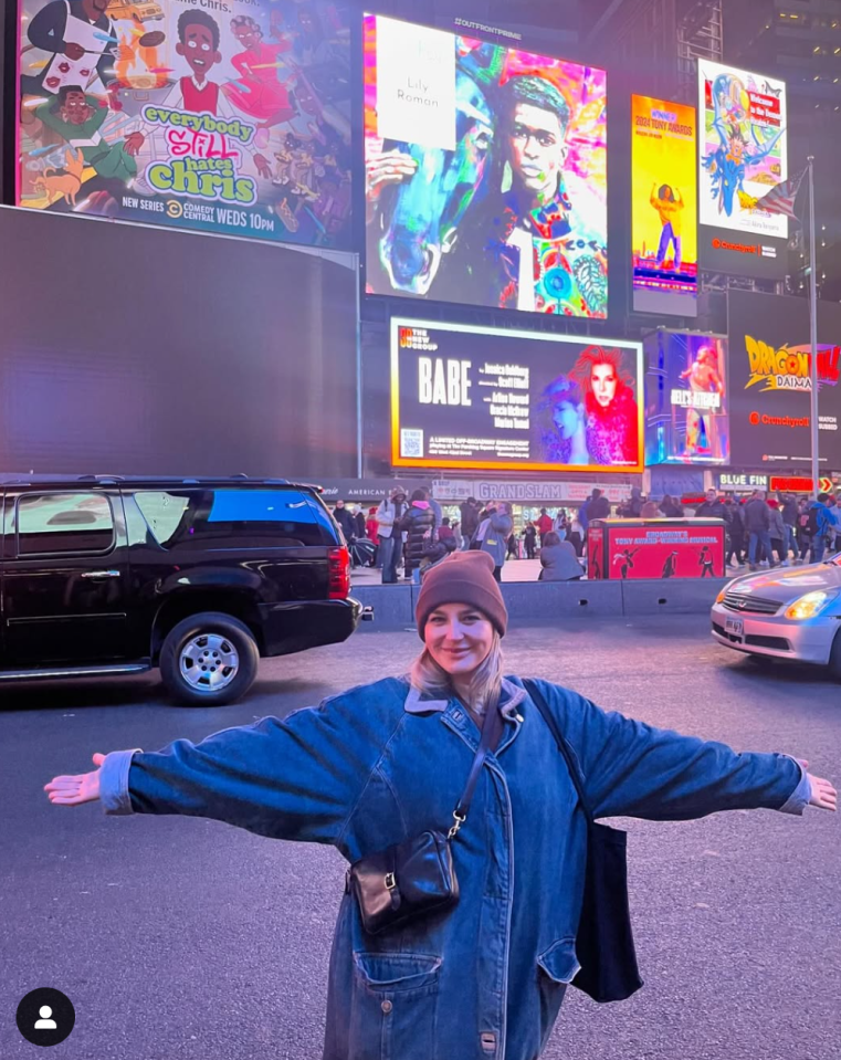 Person in Times Square with arms outstretched in front of billboards.