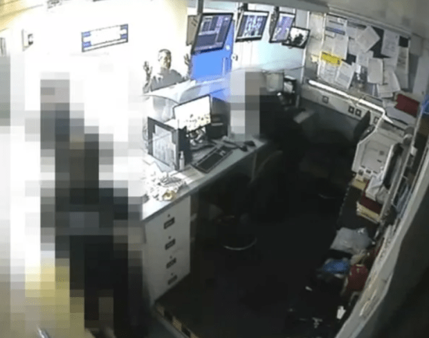 Security camera footage of a man at a desk in a cluttered office.