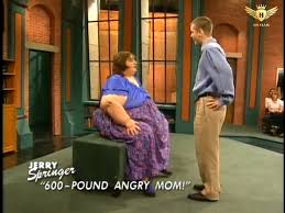 A large woman sits on a small platform on the Jerry Springer show.  A man stands facing her.