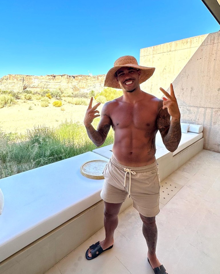 Justin Kluivert making peace signs, wearing a straw hat and beige shorts, on a patio overlooking a desert landscape.