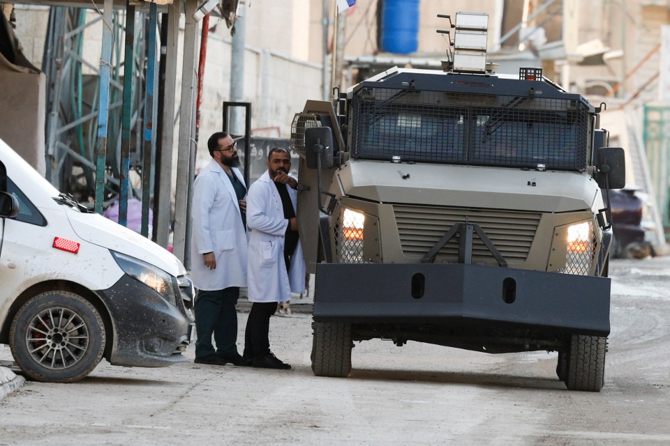 Israeli military vehicle on a street in Jenin, West Bank, with two people in medical scrubs nearby.