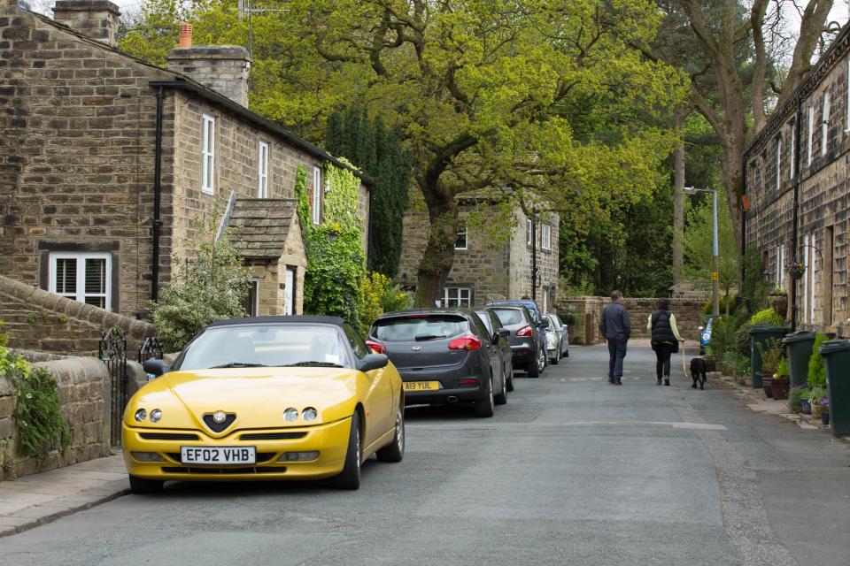 A yellow convertible car parked on a street in a village, with stone houses and people walking a dog.