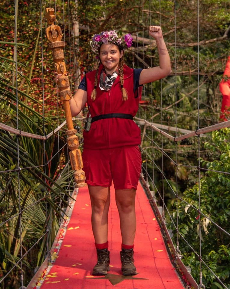 Jacqueline Jossa, winner of I'm a Celebrity... Get Me Out of Here!, holds a ceremonial staff and celebrates on a red carpet bridge.