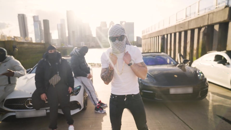 Screengrab of a music video showing a man in a balaclava and gold chains standing between two luxury cars.