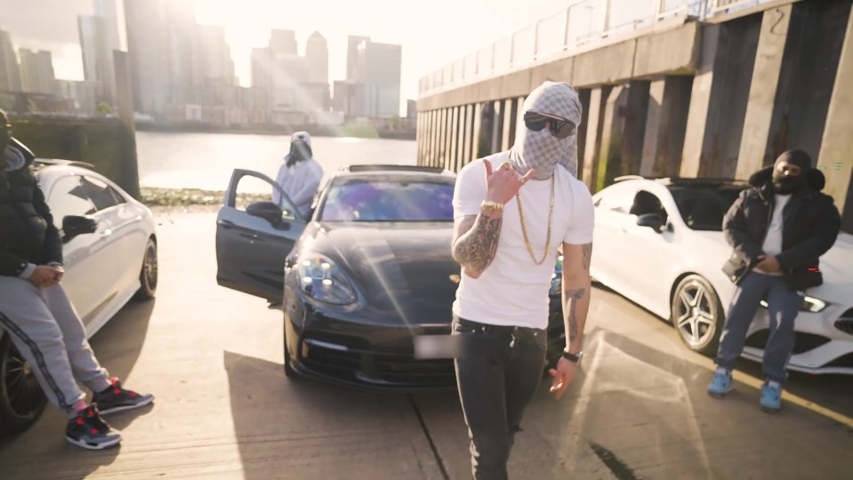 Screengrab of a music video showing a man in a balaclava and white shirt standing between two luxury cars, with other people in the background.