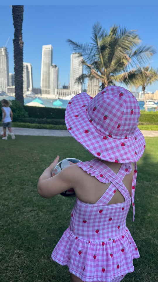 Toddler in pink strawberry swimsuit and hat holding a ball in front of Dubai skyline.