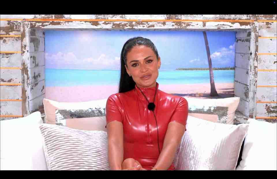 Kaz Crossley in a red dress, talking.  Beach backdrop.