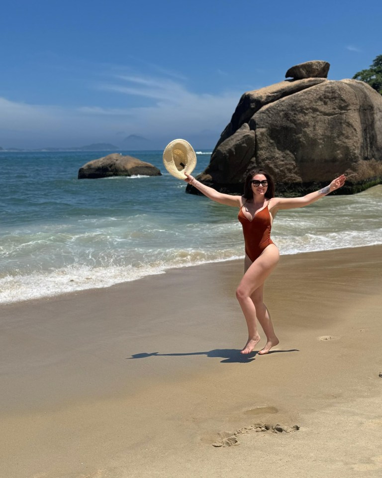 Woman in a swimsuit on a beach holding a sun hat.