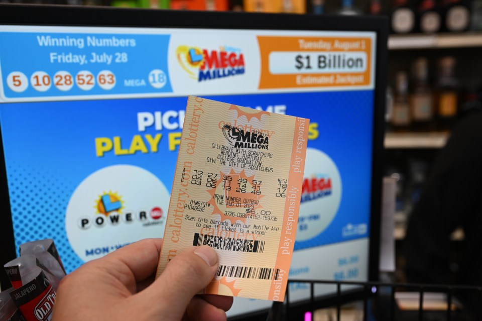 Hand holding a Mega Millions lottery ticket in front of a lottery terminal showing a $1 billion jackpot.