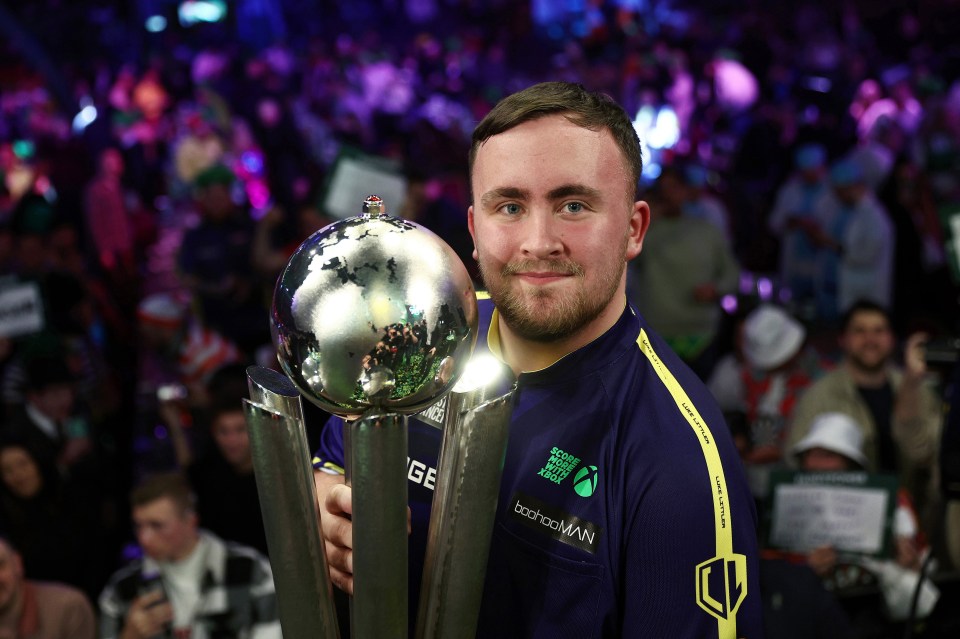 Luke Littler holding the Sid Waddell Trophy after winning a darts championship.