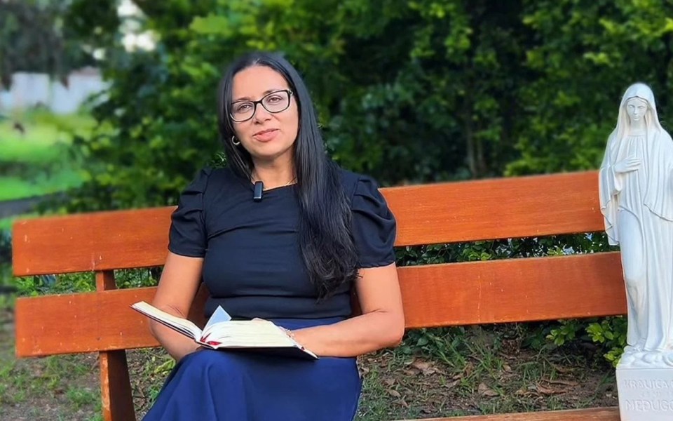 Woman sitting on a park bench holding a book, with a statue of the Virgin Mary beside her.