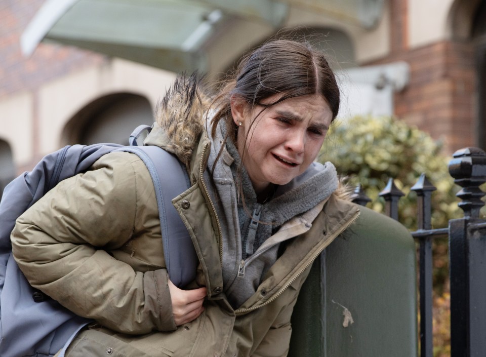 A young woman with a backpack cries while leaning against a fence.