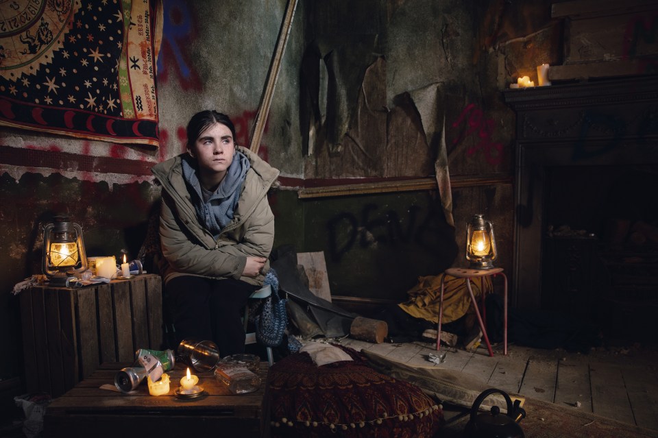 A young woman sits in a dilapidated room, illuminated by lanterns and candles.