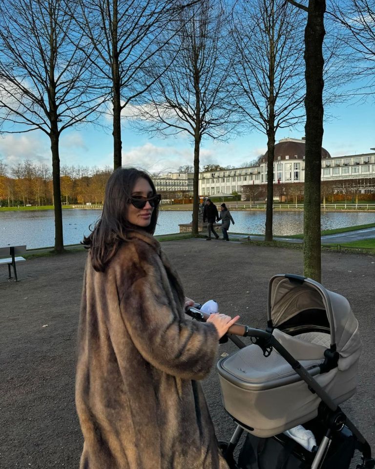 Woman in fur coat pushing a stroller by a lake.