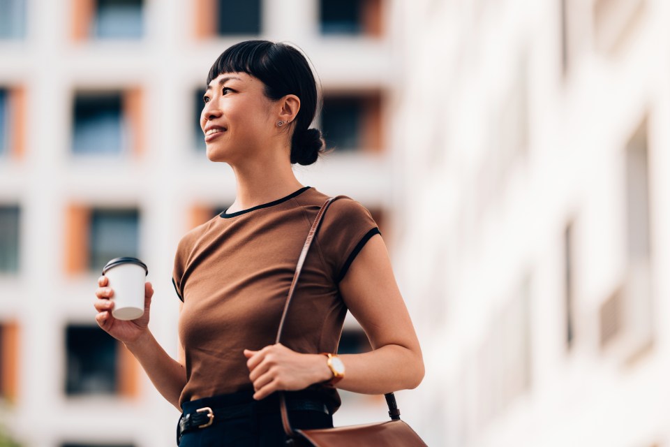 A smiling Asian businesswoman holds a coffee cup while walking in a city.