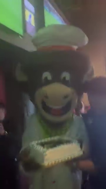 A cow mascot in a chef's hat presents a birthday cake.