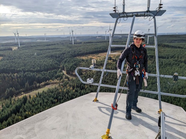 A worker stands atop a wind turbine, overlooking a vast wind farm.