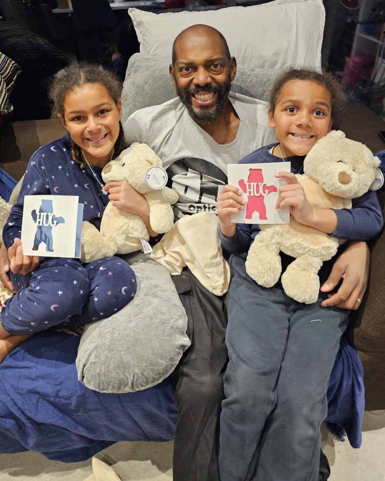 A man sits on a couch with two children, each holding a teddy bear and a card that says "HUG".