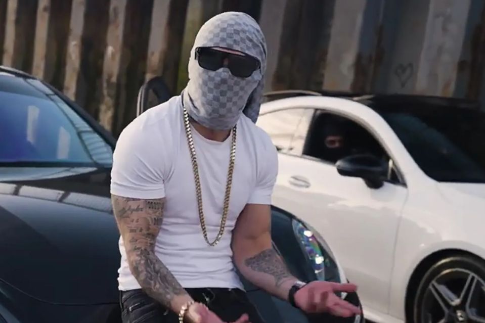 Man wearing a balaclava and gold chain sits by two cars.