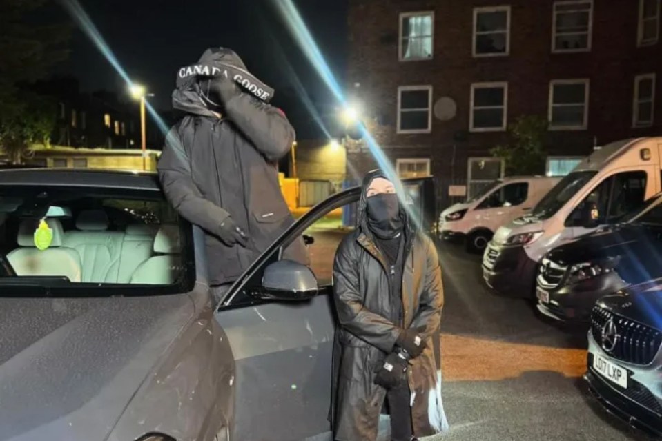 Two people in dark clothing and face coverings stand by a car at night.